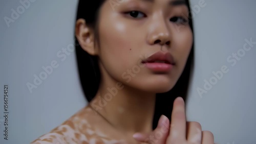 A portrait of a young Asian woman with the skin condition vitiligo. She turns from showing her back to face the camera, her expression confident and serene in a minimalist studio setting.