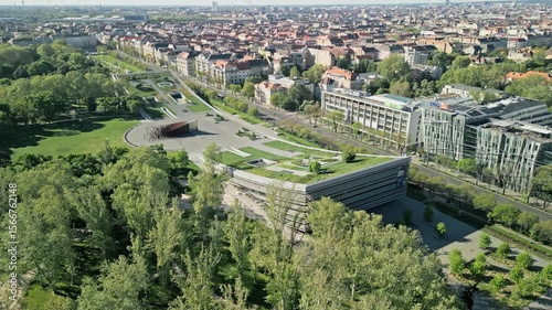 Side arc drone video keeping the Museum of Ethnography in central focus, revealing its modern curved design with Kunsthalle, City Park, and Puskás Aréna in the shifting background.  