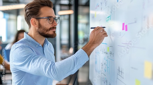A focused man in glasses writes on a whiteboard covered with diagrams and notes in a modern office setting.