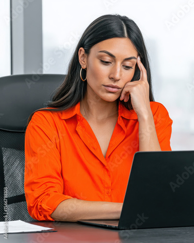 Young woman working on laptop in office wearing orange shirt with thoughtful expression