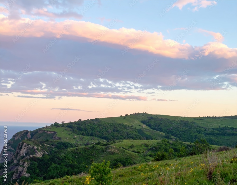 Obraz premium Lush green hills meet ocean under a pastel, cloud-strewn sky