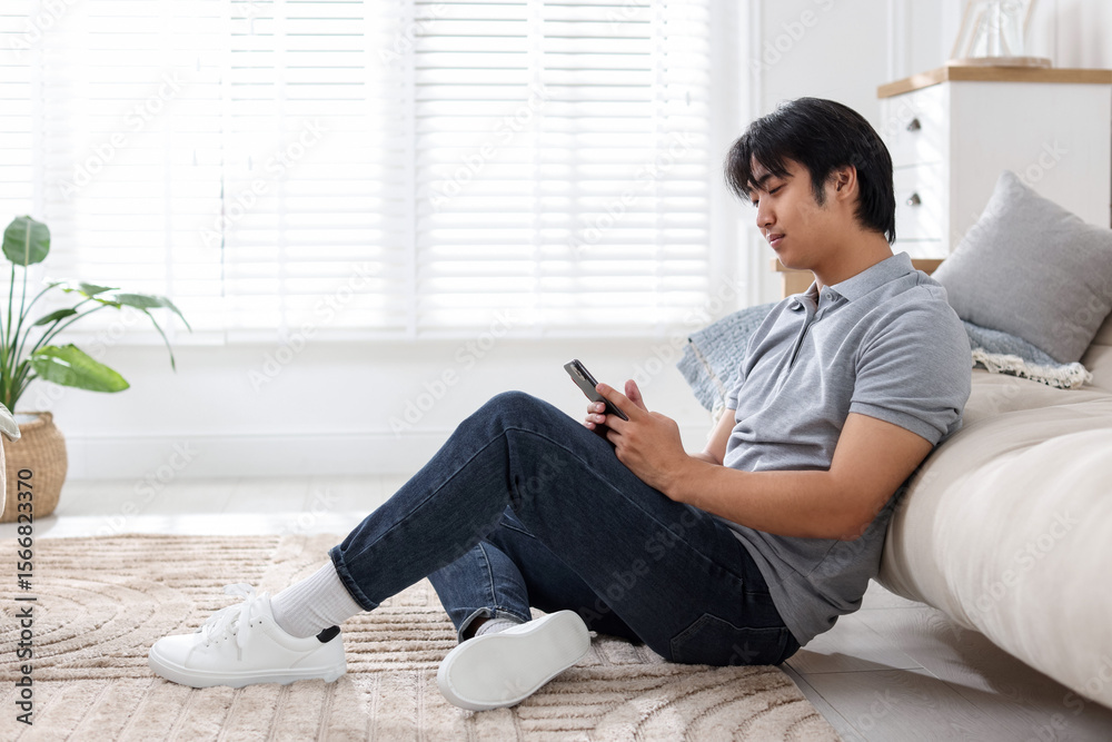 Obraz premium Handsome Asian man with smartphone on floor at home
