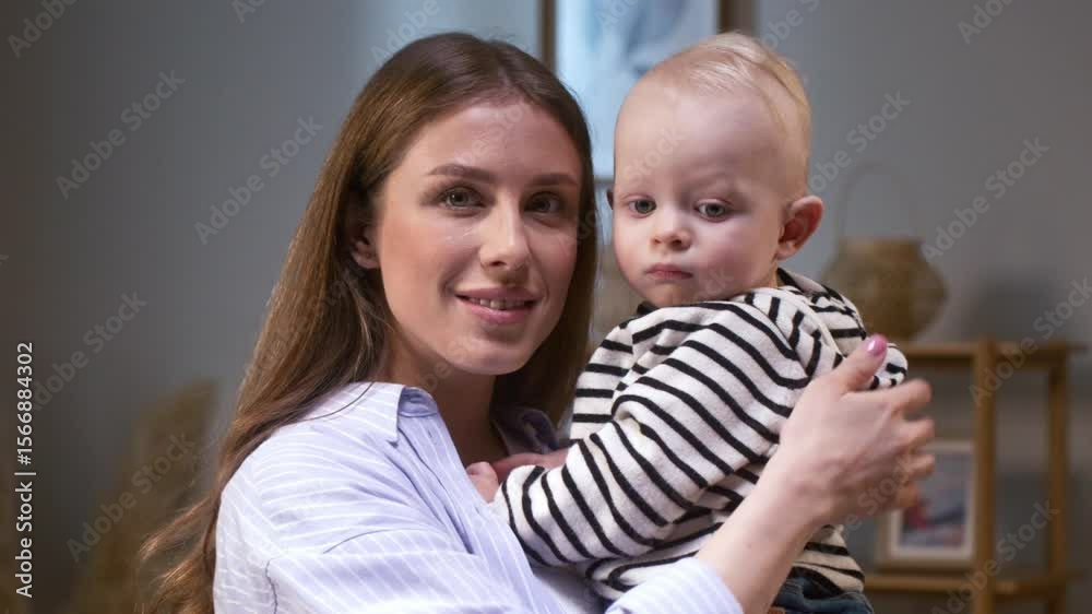 Charming woman with loose brown hair holding silent baby near bookshelf in soft daylight. Speaking gently or singing lullaby. Comforting child or enjoying peaceful moment together at home. Charming woman with loose brown hair holding silent baby near bookshelf in soft daylight. Speaking gently or singing lullaby. Comforting child or enjoying peaceful moment together at home.