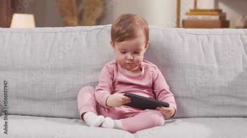 Focused toddler dressed in pink sitting on couch holding smartphone with both hands. Staring at screen and staying fully engaged. Watching animated video or tapping through playful app alone.