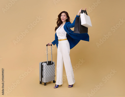 Happy young elegant Asian business woman travel drag luggage and shopping bag isolated on nude color background. Tourist girl having cheerful holiday trip concept, Full body