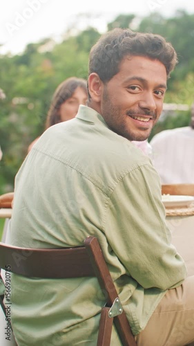 Charming man in green shirt turning around and smiling warmly while sitting at festive table. Enjoying summer dinner with friends. Relaxed outdoor setting during celebration scene.