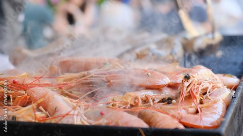 King prawns sizzle on a street grill in an open-air restaurant. A chef simultaneously cooks seafood, sausages, and meat steaks on the hot grill, preparing a mixed grill platter outdoors.
