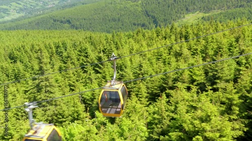 A closeup aerial tracking shot shows a yellow gondola cabin moving along a mountain cable above a dense evergreen forest and distant valley under bright summer sunlight, highlighting scenic alpine tra