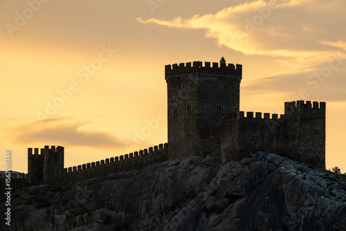 Castle Genoese Sunset Silhouette of a medieval fortress with towers and battlements on a rocky hill, against a warm sunset sky with light clouds.