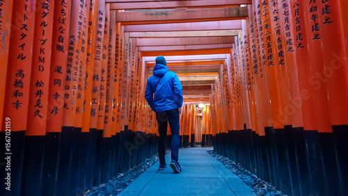A solitary man wearing a blue jacket walks down the stone path under the iconic endless rows of illuminated red torii gates at Fushimi Inari Shrine, a sacred destination in Kyoto, Japan.