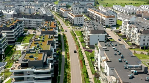 This aerial drone footage shows a smooth fly over suburban apartment buildings with green roofs, landscaped streets and tree-lined sidewalks under summer sunshine, showcasing architectural design and 