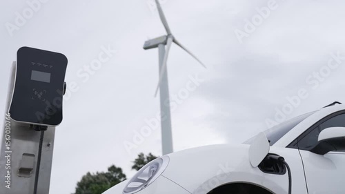 Progressive future energy infrastructure concept of electric vehicle being charged at charging station powered by green and renewable energy from a wind turbine in order to preserve the environment.