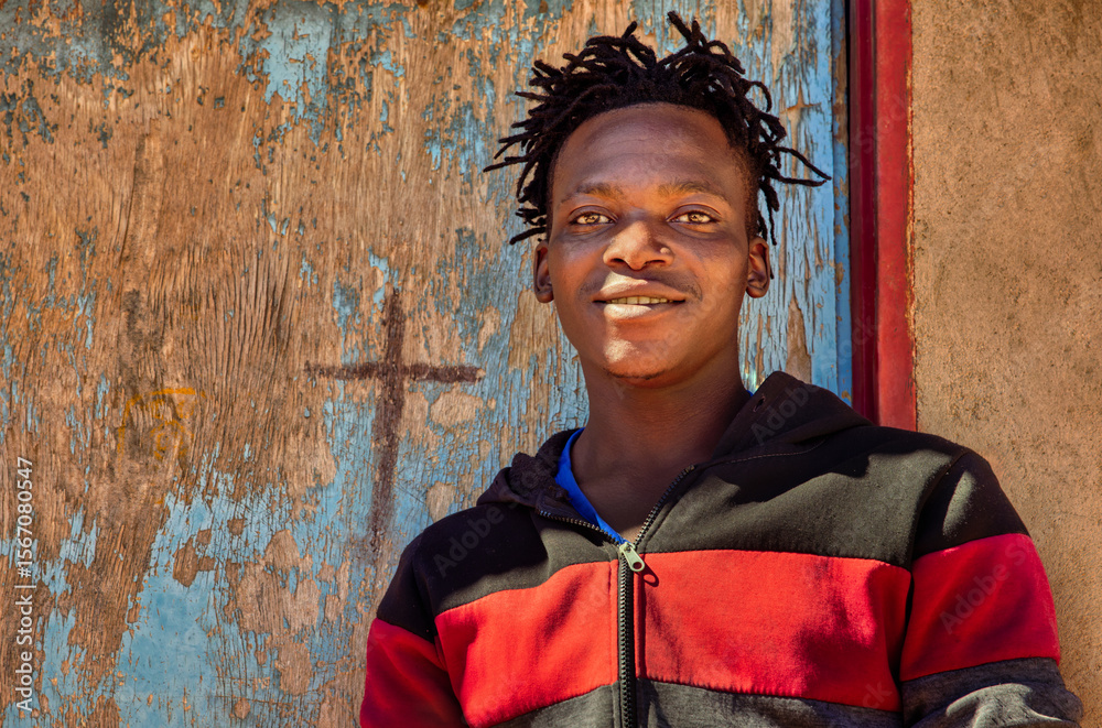 Fototapeta premium africa village, african smiling single young man with dreadlocks braids , in front of the house in the yard, blue door,