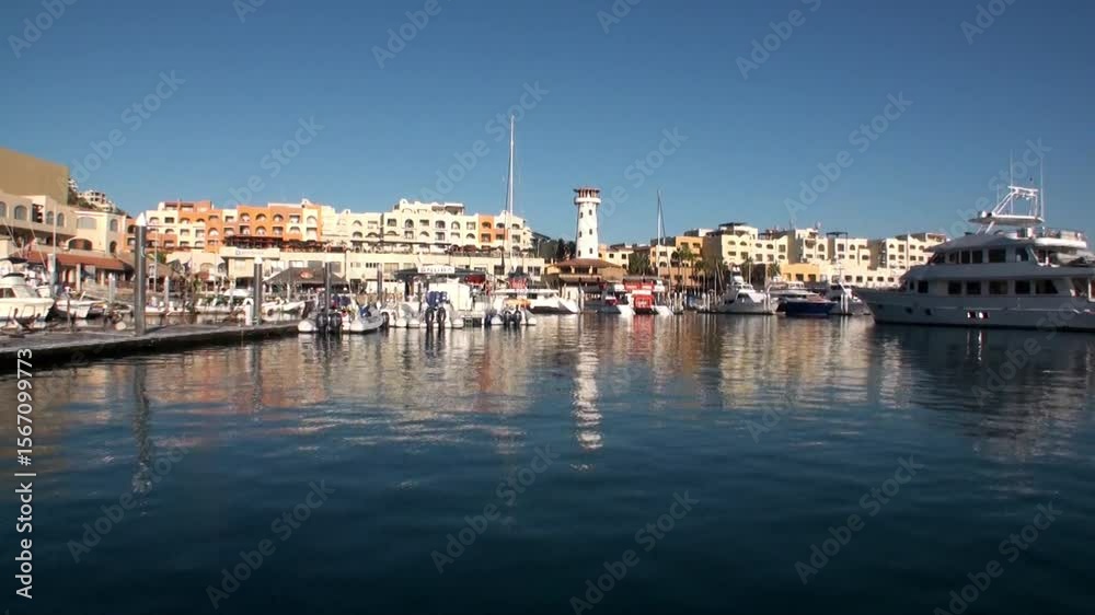 Boats line the docks in Cabo San Lucas during the day. The water reflects the sky and the structures around the Puerto Paraiso Marina in Mexico, providing a calm and serene feel.