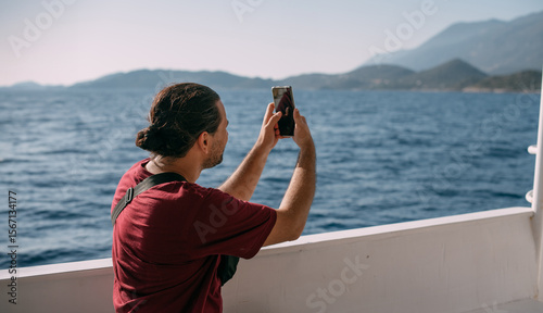 A young, contented man, a tourist rides a boat to the sea, takes pictures on the phone, enjoys a sunny day.