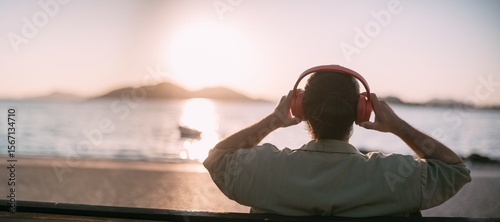 Portrait from the back of a young man in bright big headphones by the sea at sunset. A handsome guy listens to music on the ocean in the rays of the sun, with his back to the camera