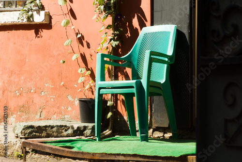 Fototapeta Naklejka Na Ścianę i Meble -  Urban street scene with two green plastic chairs stacked near a red weathered wall in bright sunlight. A small potted plant and climbing vines add charm to this quiet outdoor corner