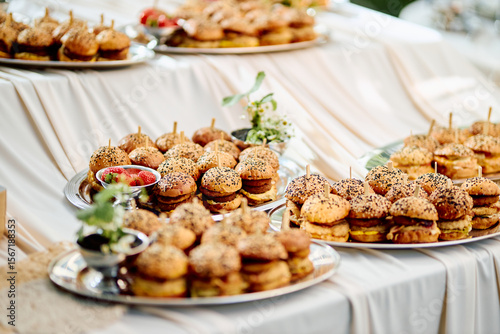 Outdoor mini sandwich platter with sesame seeds and fresh herbs, elegantly arranged on silver trays on a flowing white tablecloth, evoking a festive summer garden event