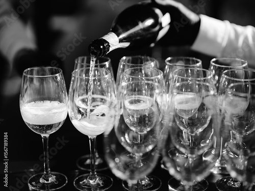 Black and white close-up of a wine server pouring sparkling wine into a row of glasses, capturing a moment of celebration or special event with reflections and focus on the bubbling drink