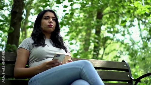 Young Woman Sits on Park Bench Holding Her Phone in Natural Sunlight With Green Trees and Casual Outfit