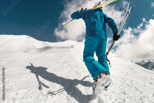 Back view of a male skier in a blue suit with skis and poles in his hands climbing up a snowy slope, bottom view