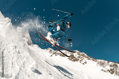 A skier performs a risky trick with a high jump, sliding down the mountain off-piste, with cloud of snow around him