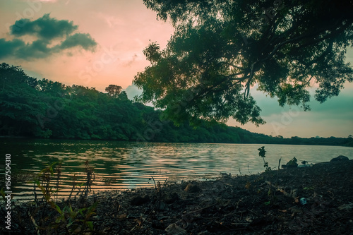 Peaceful river view surrounded by lush green trees at sunset, with calm water reflecting warm sky tones. Perfect for nature themed visuals, eco travel, and tranquil outdoor scenes
