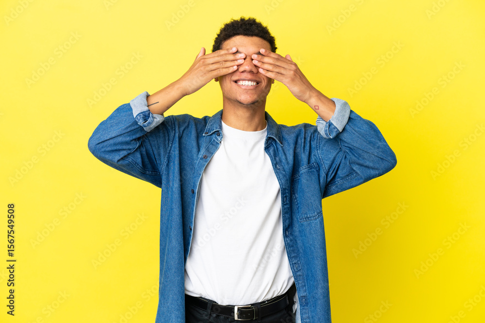 Fototapeta premium Young African American man isolated on yellow background covering eyes by hands and smiling