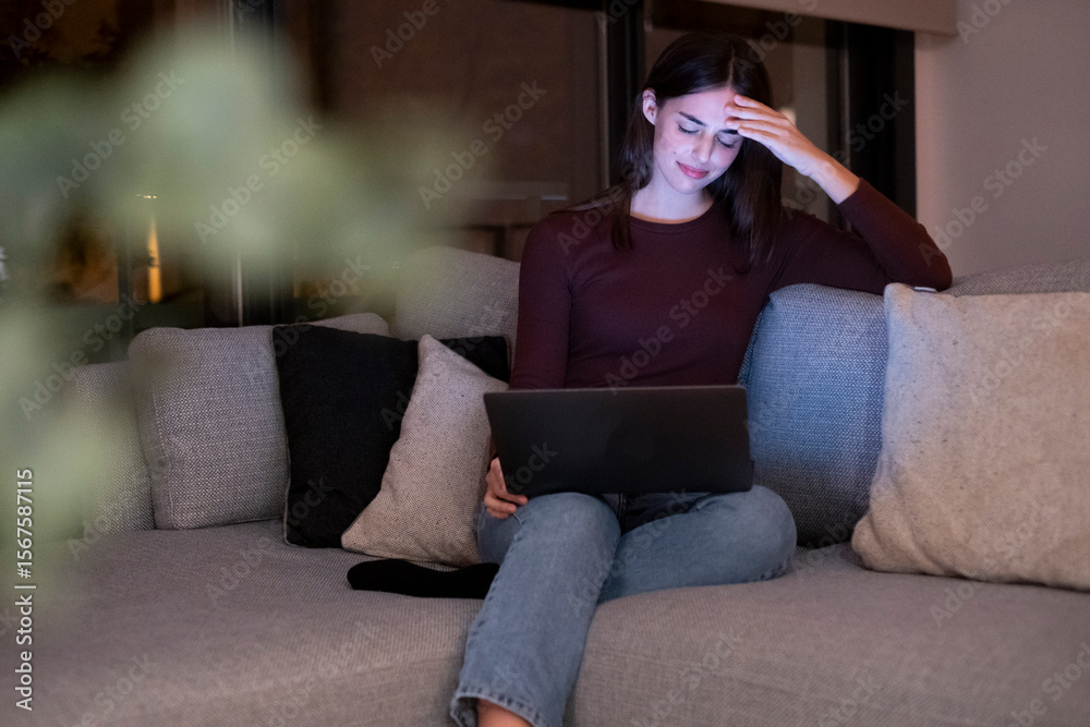 Obraz premium Young woman alone at home working late at night on her laptop. Freelancing, studying, chatting online or trying to meet a deadline—an intimate and real portrayal of modern life after dark