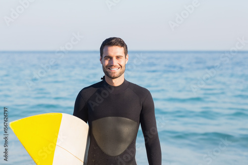 Man wearing black wetsuit standing on sandy beach holding yellow-white surfboard by ocean