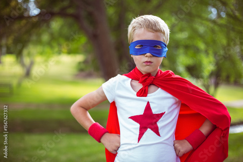 Child standing confidently in grassy park wearing red cape and wristbands with blue eye mask