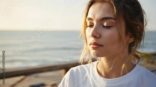 A young Caucasian woman with light brown hair is meditating by the beach at sunset. She has her eyes closed and a serene expression on her face.