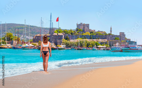 Fototapeta Naklejka Na Ścianę i Meble -  Beautiful girl in black swimsuit walks on a sandy beach -  Panoramic view of Saint Peter Castle (Bodrum castle) and marina - Bodrum, Turkey