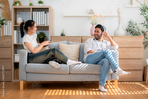Happy young asian couple relax sitting on sofa smiling and enjoy a cheerful conversation in cozy living room while man listening music with headphones and woman using smartphone at home.