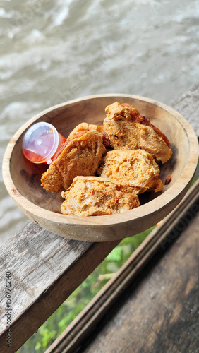 Tahu walik. Crispy browned tofu walik served in a natural wooden bowl with spicy chili sauce, with a blurry river flowing in the background. Menu at an outdoor cafe.
