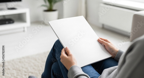 Person holding blank white magazine mockup in bright modern living room. Space for design or text.