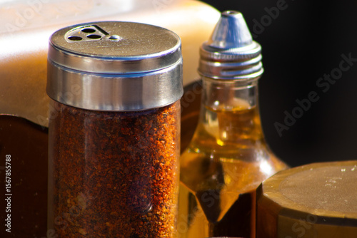 A close-up shot of various seasoning bottles and a jar of pickled peppers resting on a rustic outdoor table, bathed in warm sunlight