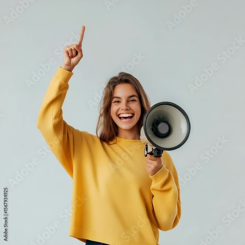 A young woman in a yellow sweater smiles happily, holding a loudspeaker and pointing her index finger up, symbolizing enthusiasm, engagement, and the power of communication.
