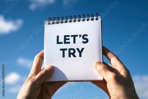 Motivation shown as a person holds up a notepad with the text Let's Try against a bright blue sky for inspiration.