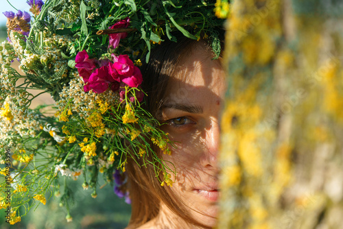 A girl of Slavic appearance with a wreath of wild flowers on her head looks out from behind a tree