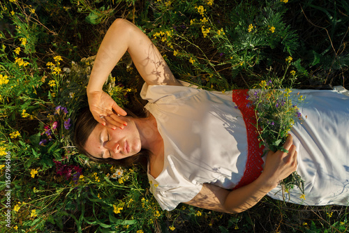 A girl in a white dress, of Slavic appearance, with a bouquet and a wreath of wild flowers on her head, lies in a meadow.