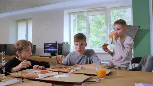 Three boys are seated at a table in a modern classroom, sharing pizza and orange juice. They are happily engaged in conversation while eating their meal.