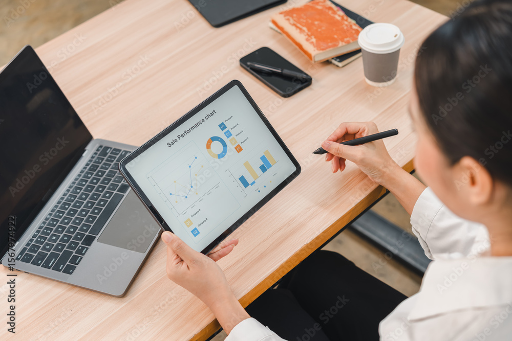 Businesswoman analyzing sales performance chart on digital tablet, working at modern office desk with laptop, notebook, smartphone, and coffee, focused and professional atmosphere Businesswoman analyzing sales performance chart on digital tablet, working at modern office desk with laptop, notebook, smartphone, and coffee, focused and professional atmosphere