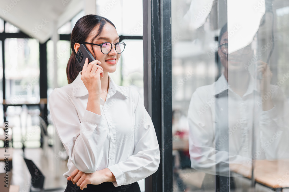 © kenchiro168 - Young Asian businesswoman with glasses standing indoors, smiling while talking on smartphone, wearing white shirt, modern office background, confident and professional mood © kenchiro168 - Young Asian businesswoman with glasses standing indoors, smiling while talking on smartphone, wearing white shirt, modern office background, confident and professional mood