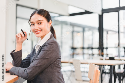Confident businesswoman Asian woman professional suit sitting at modern office desk smiling holding pen natural light workplace success positive emotion leadership career