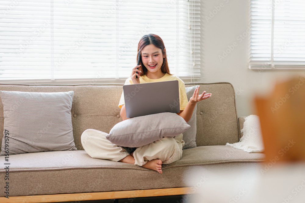 Fototapeta premium Young woman sitting on sofa with laptop, talking on phone, working from home, casual and relaxed atmosphere, natural light from window, comfortable living room