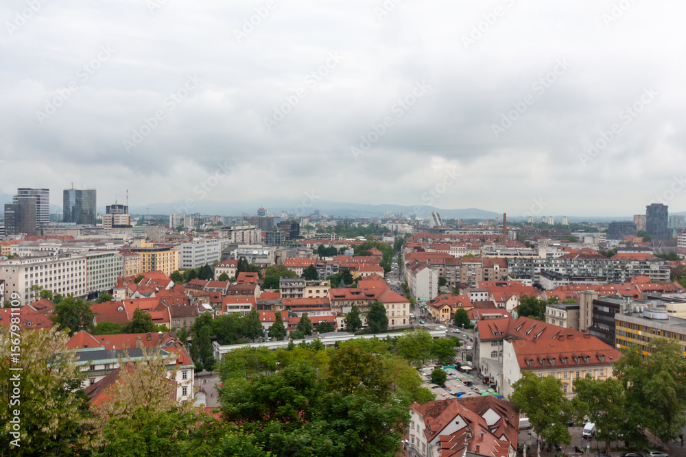 Fototapeta premium An expansive elevated view captures the cityscape of Ljubljana, on a cloudy day. Red-tiled rooftops dominate the foreground, showcasing the charming historic architecture of the old town