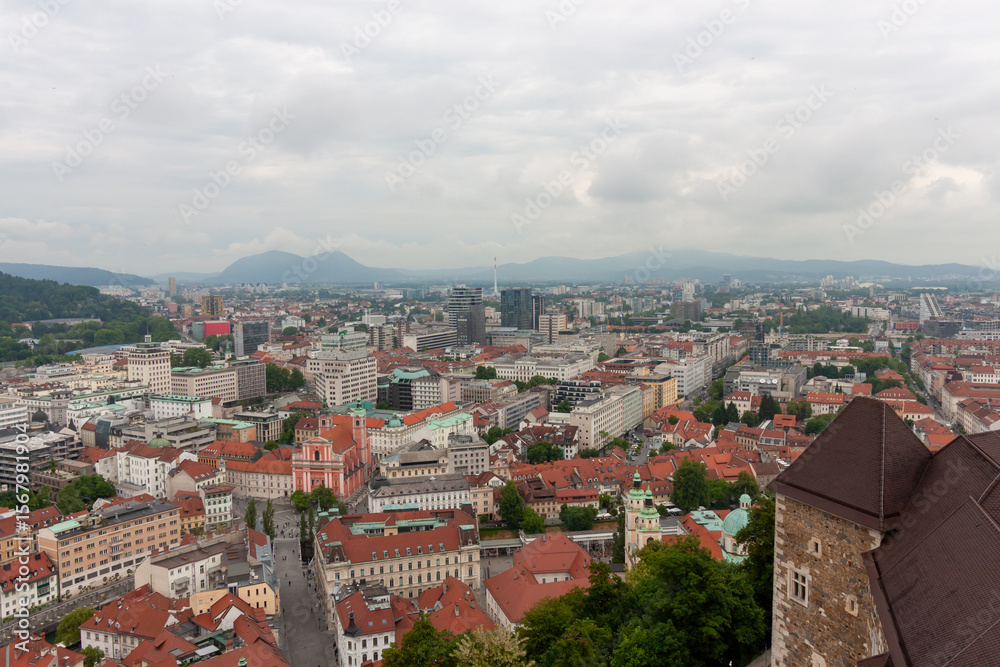 Fototapeta premium An expansive elevated view captures the cityscape of Ljubljana, on a cloudy day. Red-tiled rooftops dominate the foreground, showcasing the charming historic architecture of the old town