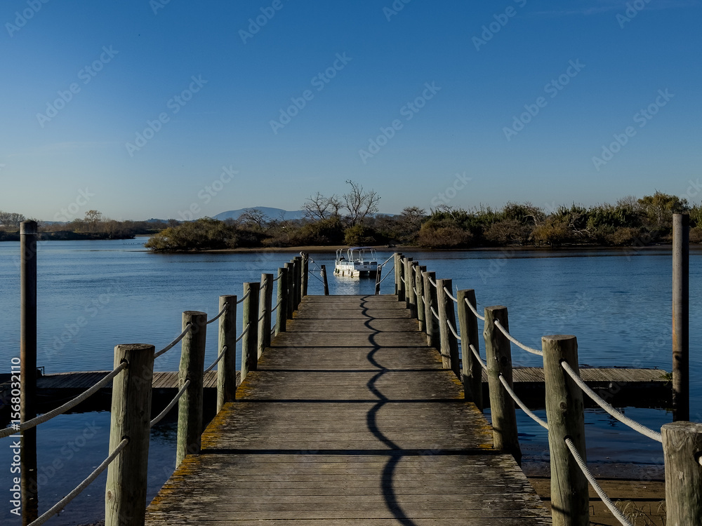 Fototapeta premium Straight wooden pier with rope railings points to a small white motorboat moored on calm water. Low brush lines the far bank and distant hills sit under a clear blue sky, giving a quiet riverside mood
