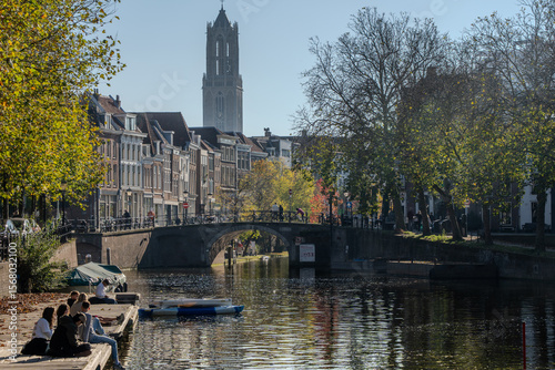 Sunny autumn view along Utrecht’s Oudegracht. People relax on the wharf as a pedal boat drifts by, an arched stone bridge spans the water, and the Dom Tower rises above historic canal-house façades. 